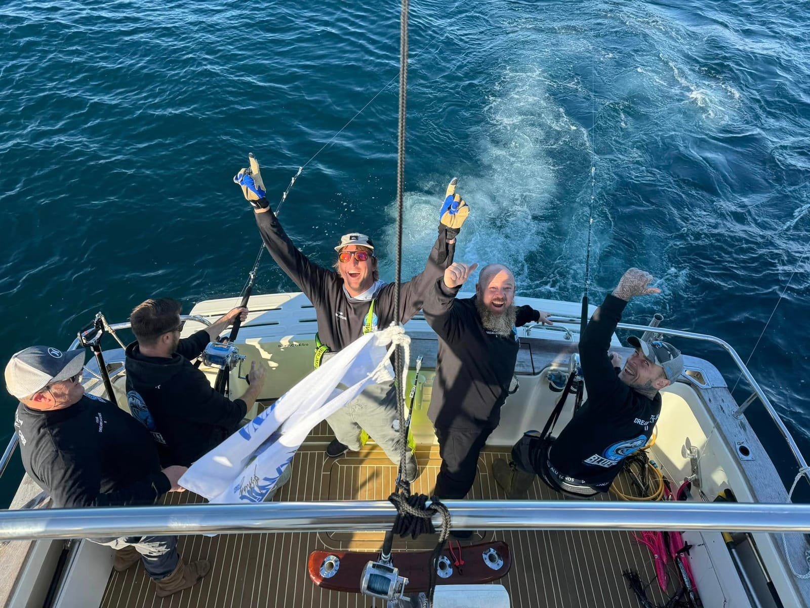 Four anglers on the deck of Dreamcatcher cheering up at the camera with the wake behind them